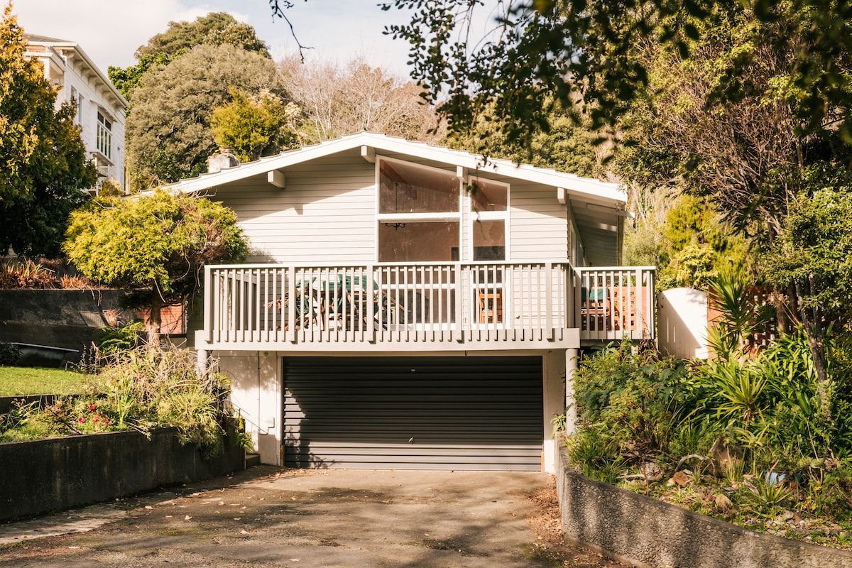 The exterior of the property features a modern house with a spacious deck surrounded by greenery. Large windows allow natural light to fill the interior, while the driveway leads to a closed garage. A serene setting is accentuated by mature trees and shrubs.