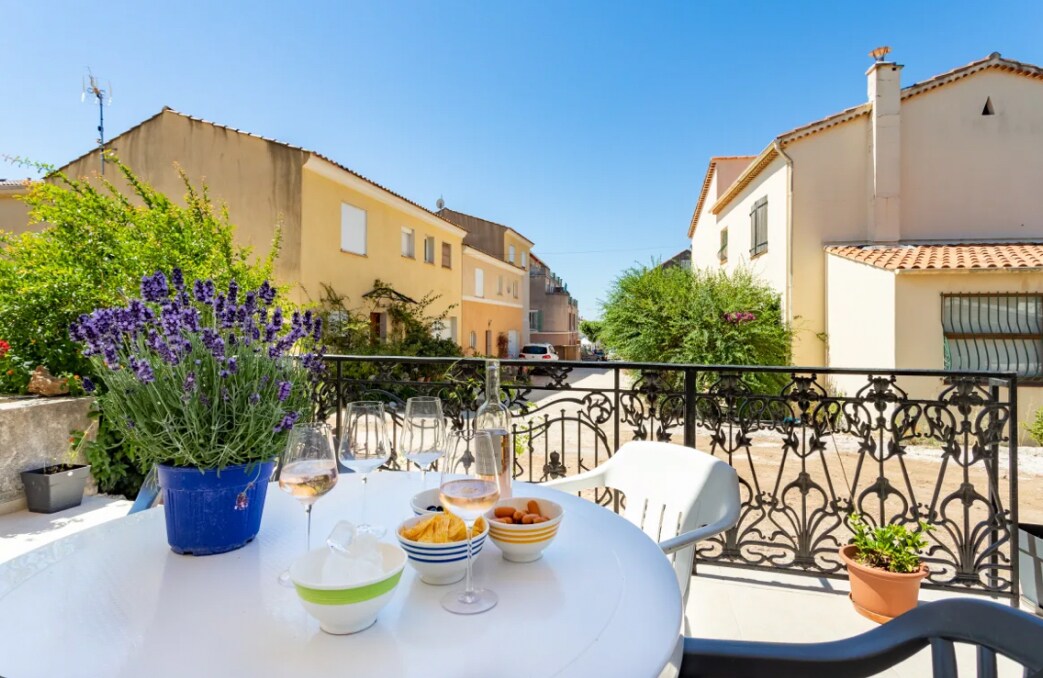 A terrace table is set with snacks and glasses of wine, surrounded by lush lavender in a blue pot. The view includes calm residential streets with pastel-colored buildings under a clear blue sky.