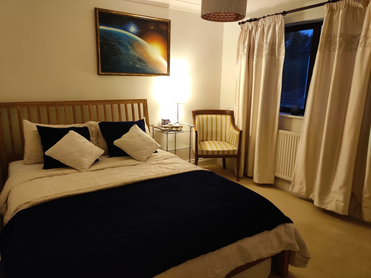 A serene bedroom is featured, with a spacious bed adorned with white and navy bedding. An artistic depiction of Earth hangs on the wall above, while a bedside lamp casts a warm glow. A striped chair and curtained window complement the neutral decor.