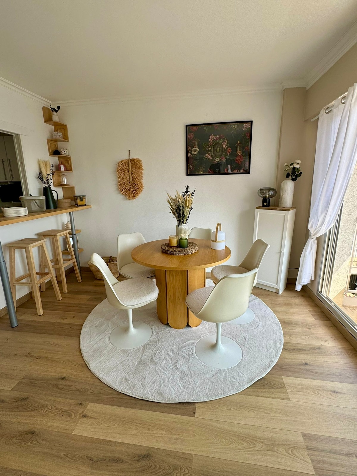 A circular wooden dining table is surrounded by four white chairs, placed on a round area rug. A small vase with dried flowers adorns the center. Natural light filters in through adjacent glass doors, enhancing the light wood flooring and creating a warm ambiance.