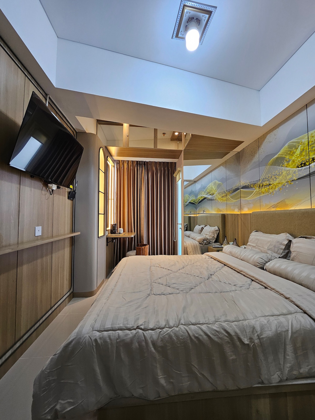 A spacious studio is presented with a king-size bed and an aesthetic mirror above a wooden shelf. Natural light filters through large curtains beside a wall featuring decorative artwork. A flat-screen TV is mounted for entertainment, enhancing the modern decor.