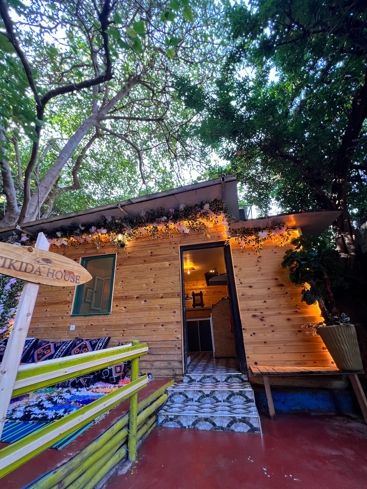 A wooden cabin with an inviting exterior is framed by lush greenery. String lights and flower accents adorn the facade, illuminating the entryway. Colorful seating is arranged on the porch, while a patterned tile pathway leads to the door, creating a welcoming entrance.