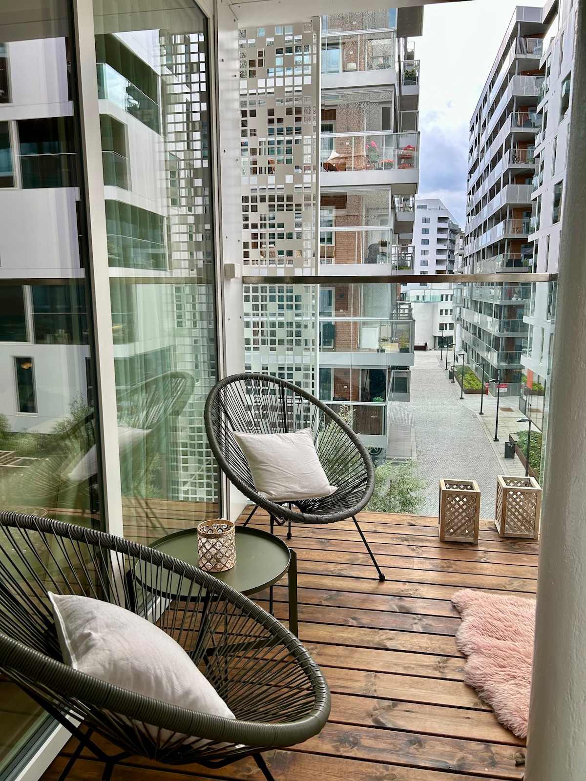 A private balcony features two modern chairs alongside a small table. Soft pillows are placed on the chairs, while stylish lanterns sit on the wooden floor. The view includes surrounding buildings and a pathway, inviting a sense of urban tranquility.