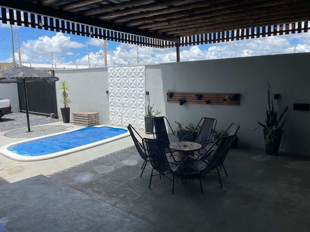 An outdoor area features a swimming pool surrounded by a concrete deck. A round table with six black chairs is positioned nearby, and decorative plants are placed around the space. The area is sheltered by a wooden roof, providing shade from the sun.