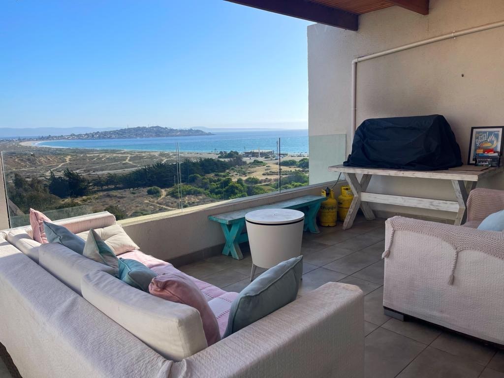 A comfortable outdoor seating area is featured, with a light-colored sofa adorned with pastel cushions. A barbecue grill is positioned on a table, alongside a small round table. The ocean and coastal landscape are visible in the background, with clear blue skies enhancing the view.
