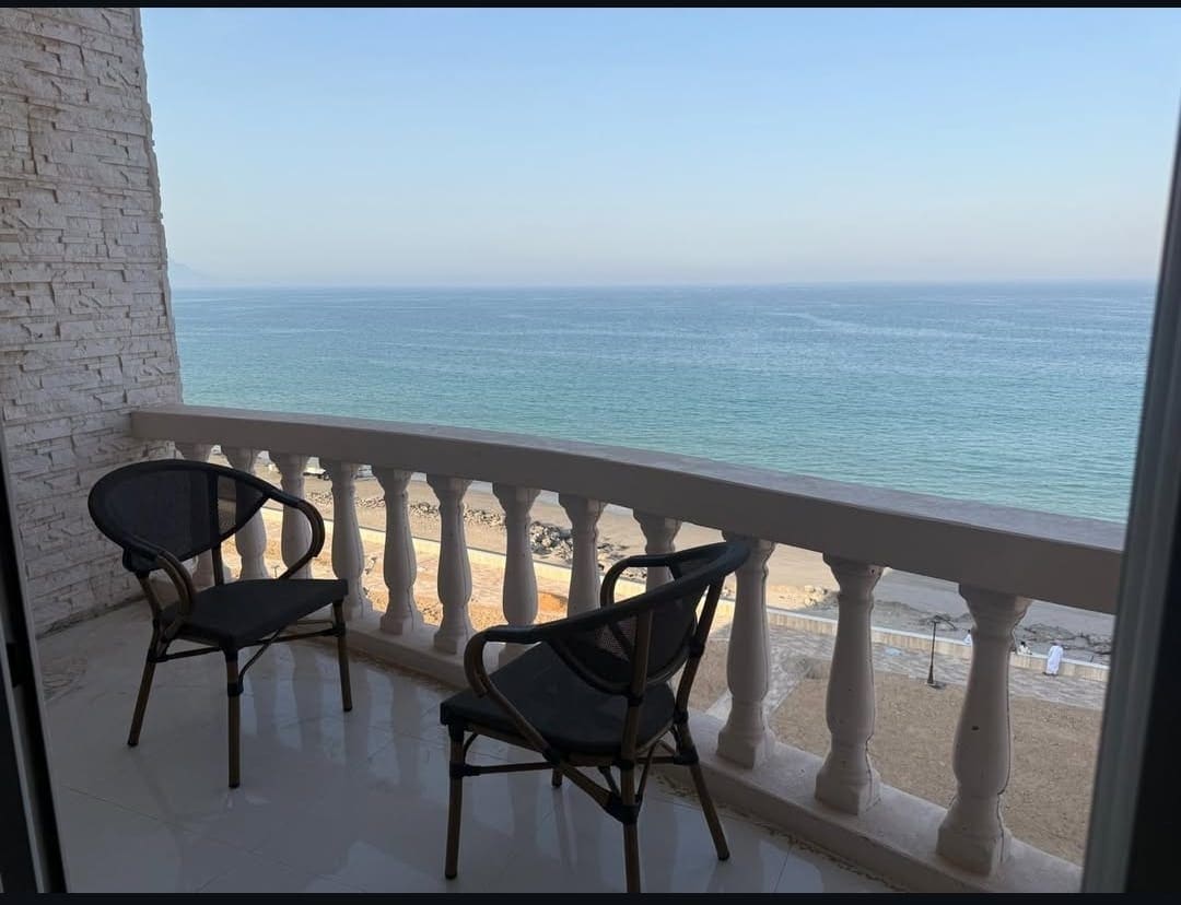 A balcony features two black chairs positioned beside a decorative railing. The clear sea view stretches out towards the horizon, showcasing gentle waves and sandy beach below. The tranquil atmosphere is complemented by the bright sky and calm waters.