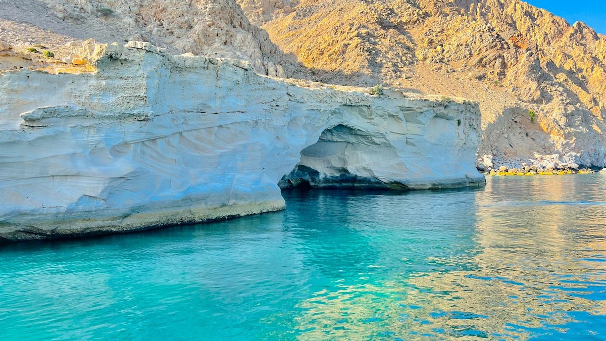 A serene coastal scene is depicted, showcasing a striking rock formation with smooth textures and a natural arch. The crystal-clear water reflects vivid shades of turquoise, while the surrounding rocky cliffs rise gently in the background, evoking a sense of tranquility.