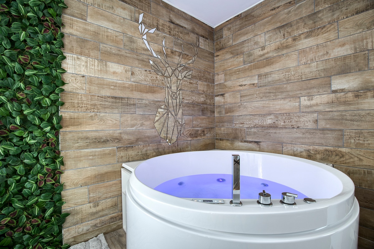 A round jacuzzi is positioned against a backdrop of textured wood-like tiles and a mirrored deer head accent. The walls are partially decorated with vibrant green foliage, creating a serene and intimate atmosphere. Soft purple lighting casts a calming glow across the water's surface.