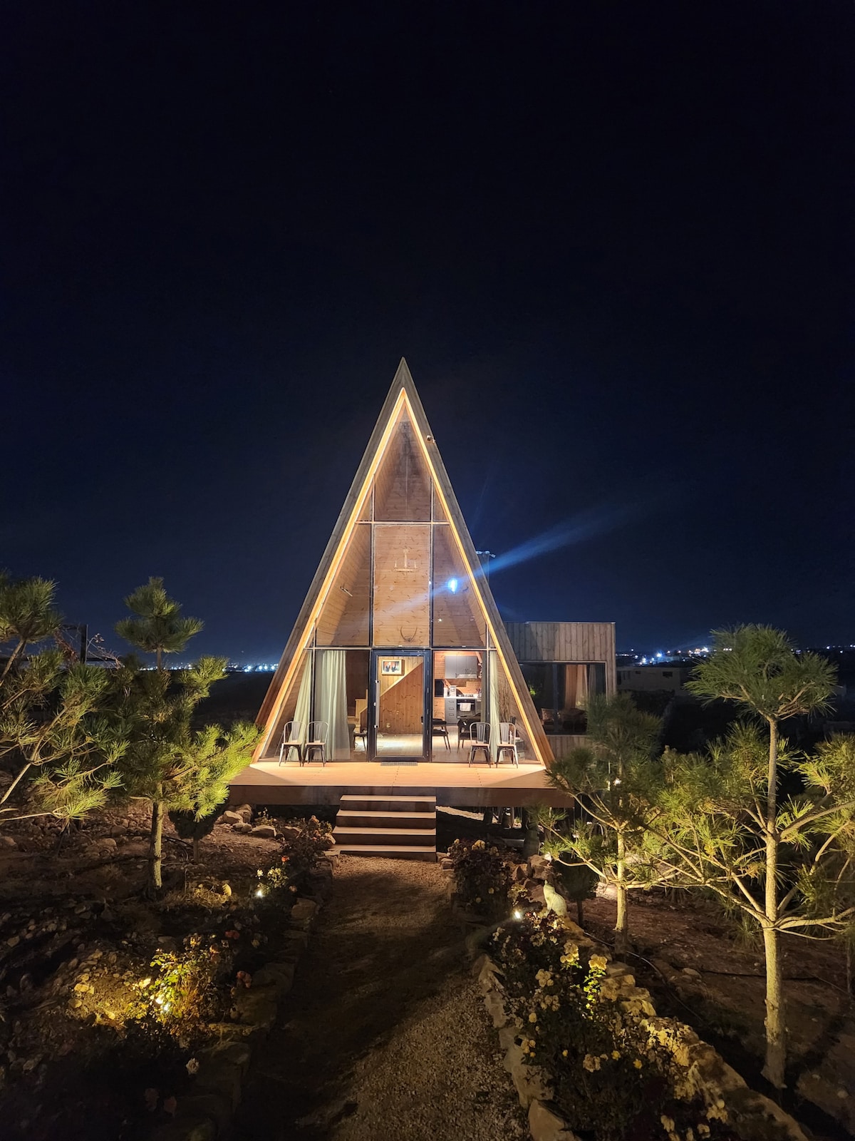 A triangular-shaped wooden house stands illuminated against the night sky, with accent lighting highlighting its architectural features. Surrounding greenery and a pathway lead to the entrance, which is flanked by two chairs on a small porch.