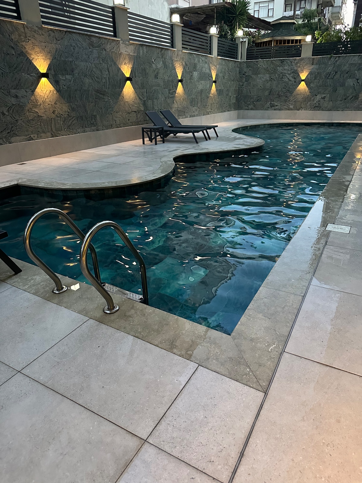A serene swimming pool is showcased, featuring smooth stone tiles and softly illuminated walls. Two lounge chairs are placed beside the water, creating a relaxing atmosphere for guests. The pool's clear water reflects light, enhancing the tranquil setting.