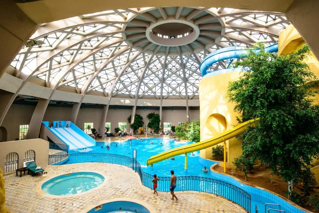 An indoor water park is showcased, featuring a large turquoise pool surrounded by bright yellow structures. A water slide curves down into the pool, with several guests enjoying the water. A smaller hot tub is positioned near the pool's edge, framed by tropical plants.