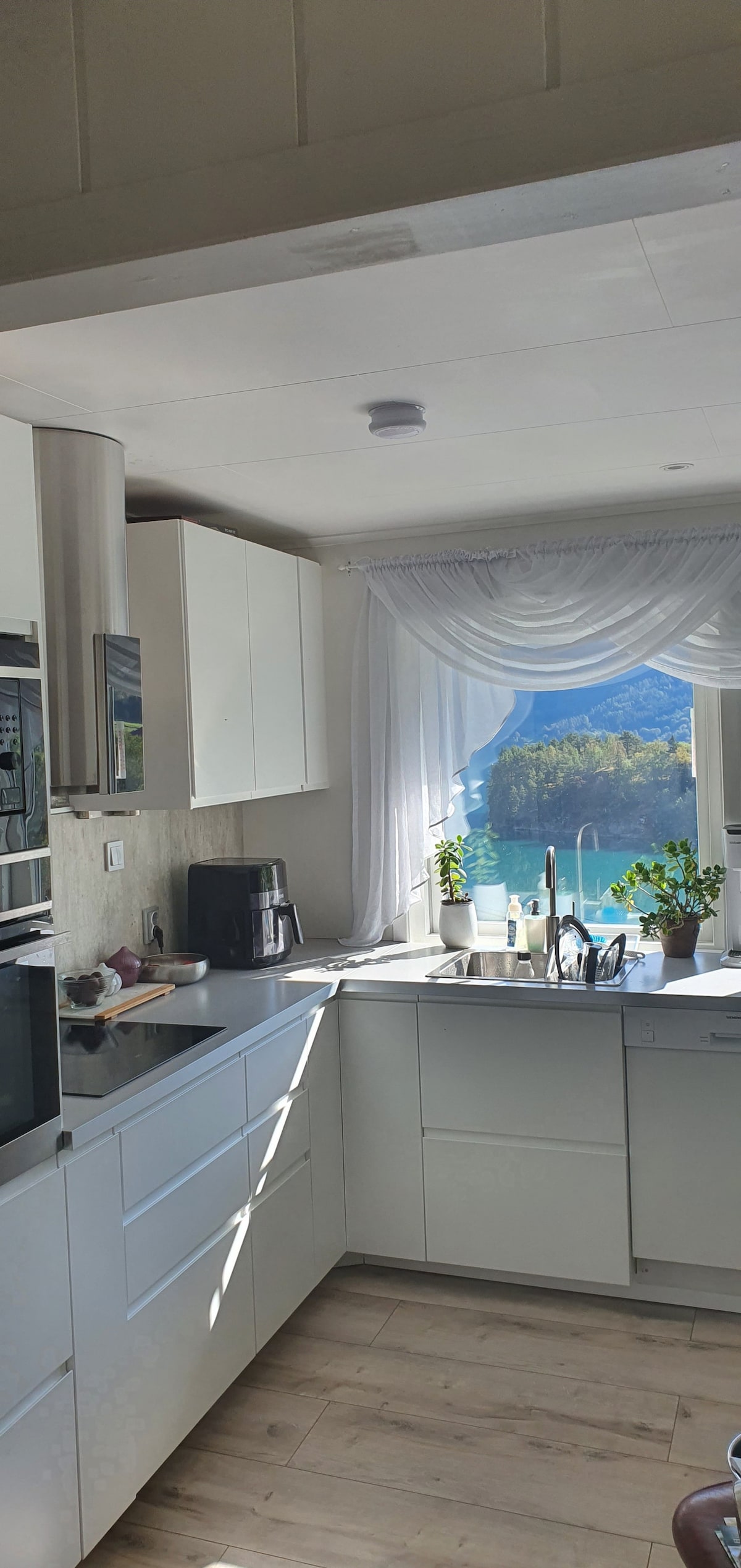 A modern kitchen is showcased with sleek white cabinetry and stainless steel appliances. Natural light pours through a large window, accentuating a scenic view of Barsnesfjorden. Potted plants are placed near the window, contributing to a fresh and inviting atmosphere.