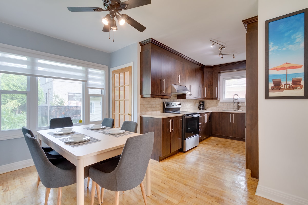 A modern kitchen is showcased, featuring dark wooden cabinetry and light-colored countertops. A dining table with four chairs is positioned nearby. Natural light enters through multiple large windows, enhancing the spacious feel of the area. A ceiling fan is present for added comfort.