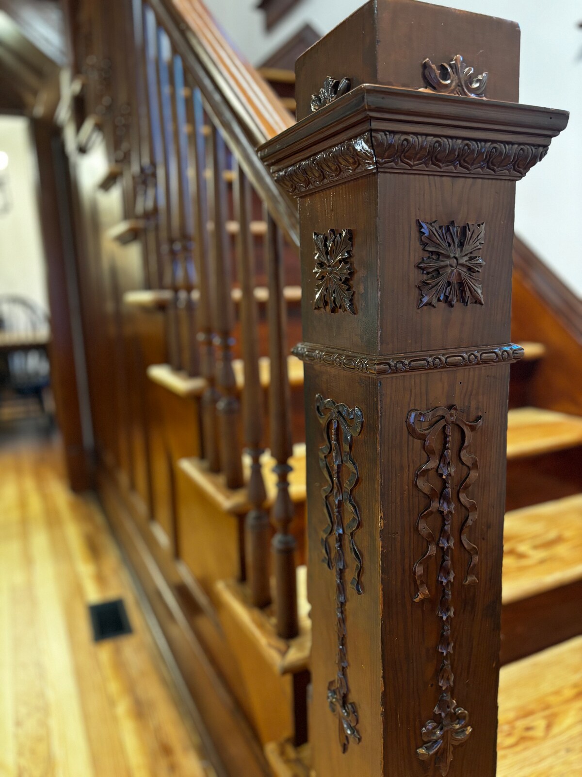A detailed close-up of a wooden staircase railing showcases intricate carvings and decorative elements. The polished banister leads upward, complemented by a series of slender spindles. Warm wooden tones reflect the craftsmanship of the historic farmhouse, while smooth stairs descend into view.