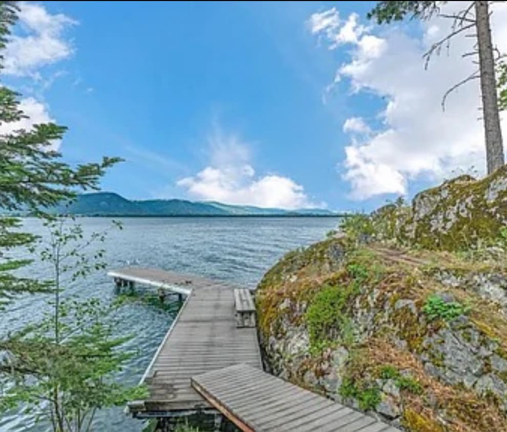 A wooden dock extends into the water, surrounded by rocky formations and lush trees. The calm lake reflects the blue sky and distant mountains, providing a serene backdrop. The area conveys a natural and tranquil setting, enhancing the waterfront experience.