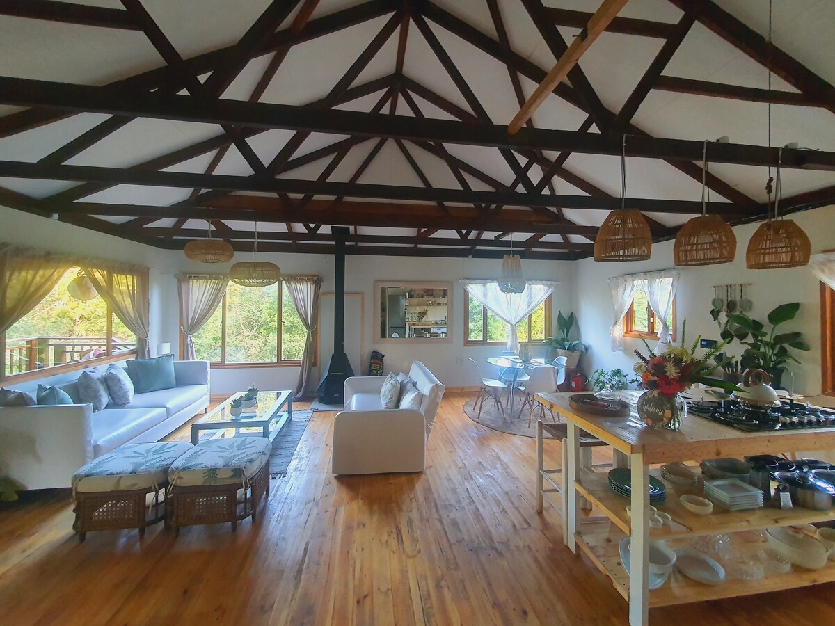 The spacious open-plan living area features exposed wooden beams and large windows that allow natural light to fill the space. A mix of seating options, including a sectional sofa and white chairs, is complemented by a dining table. An indoor fireplace is situated at the center.