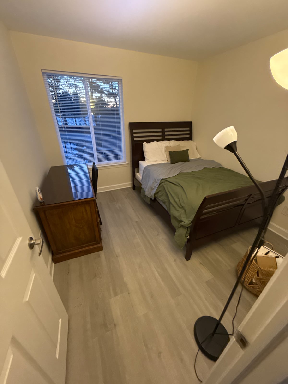 A cozy bedroom is furnished with a spacious bed layered with soft linens and a decorative pillow. A wooden dresser is positioned against the wall, and natural light enters through a large window, complemented by a simple floor lamp nearby.