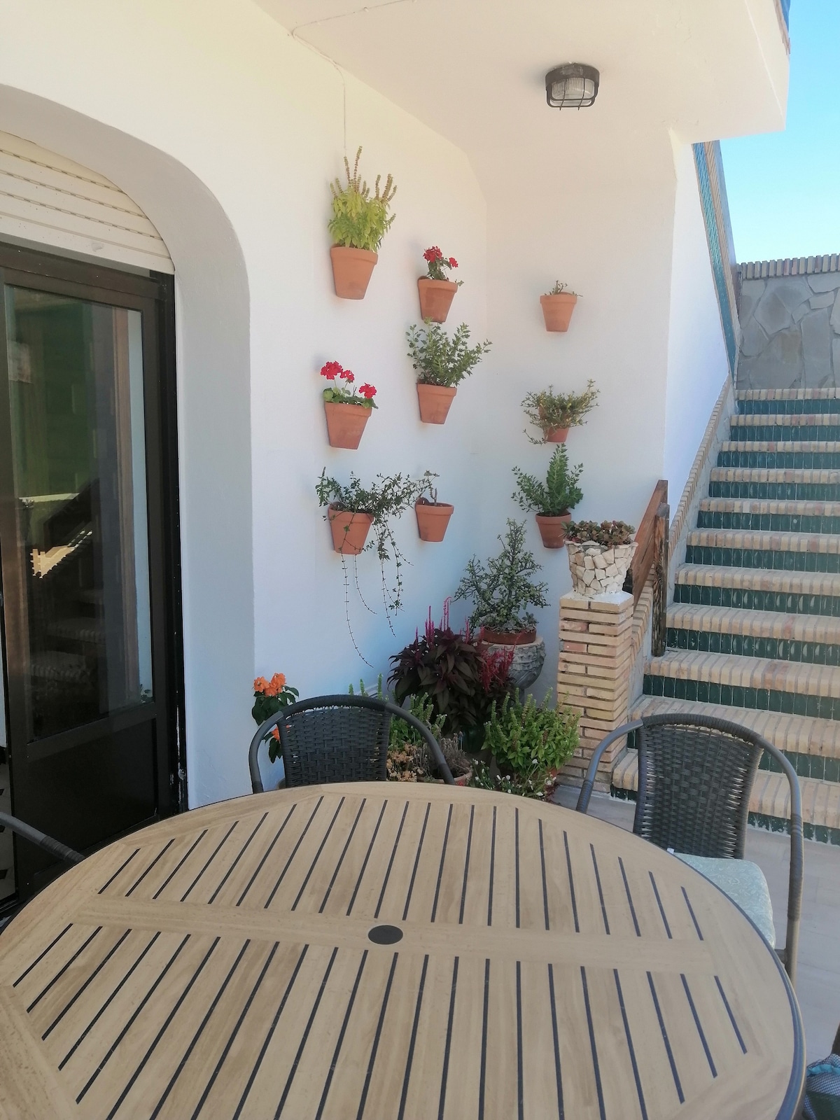 An outdoor seating area features a wooden table surrounded by four woven chairs. The wall is adorned with several potted plants, adding greenery to the space. A staircase leads up, providing access to the upper level of the property.