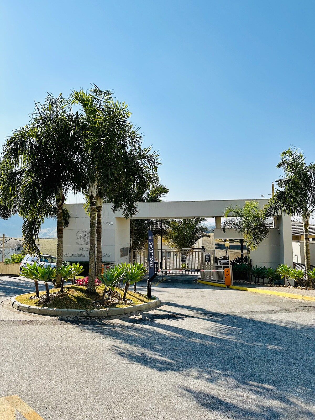 The entrance of a gated community is framed by palm trees and lush greenery. A clear blue sky forms a backdrop, while well-maintained landscaping surrounds the entrance. A security gate and signage are visible, emphasizing the safe and organized nature of the property.