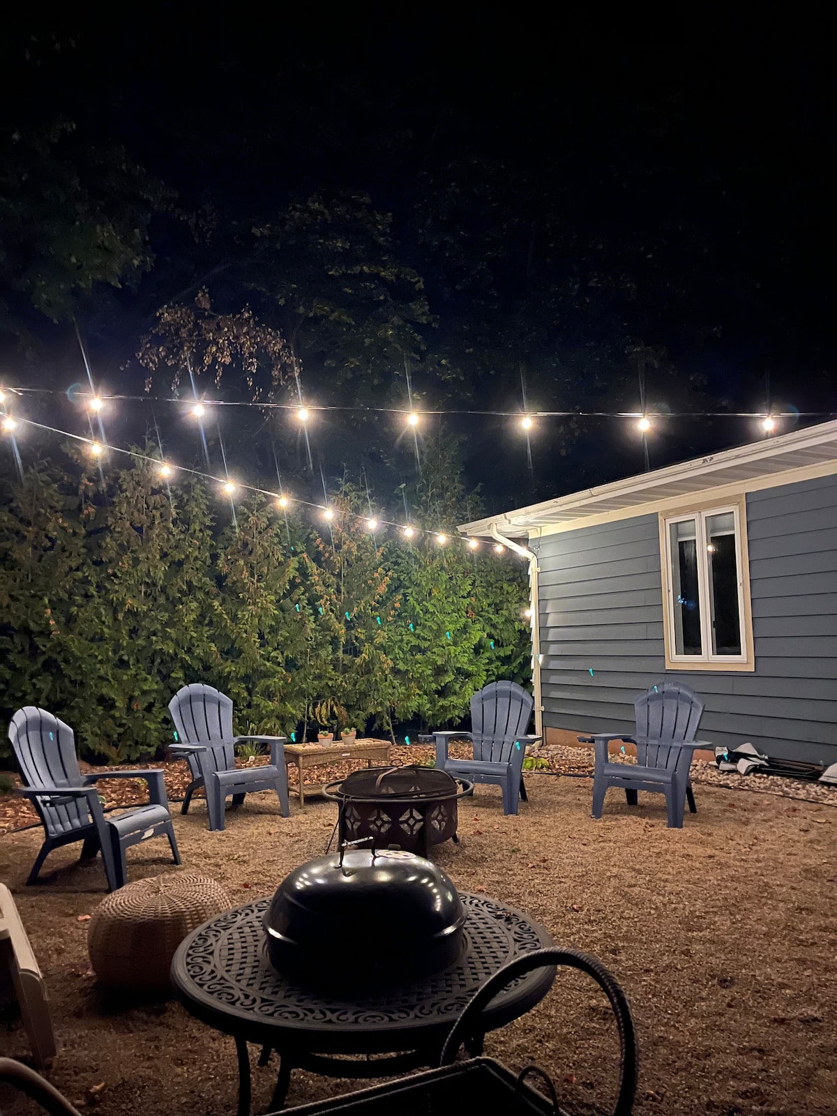 A cozy outdoor space is illuminated by string lights, creating a welcoming ambiance at night. Several blue Adirondack chairs are arranged around a fire pit, enhancing social interaction. A circular metal table with an ornate design is visible, surrounded by gravel and greenery.