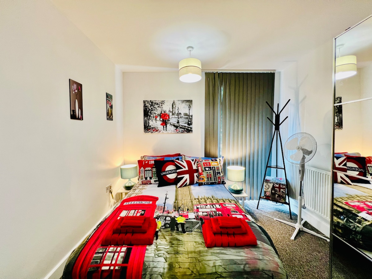 A bright bedroom features a bed with a London-themed duvet cover, accented by red towels. A mirror reflects the light from a stylish ceiling lamp, while a modern fan and a coat rack are positioned nearby. Blackout curtains offer privacy, enhancing the cozy atmosphere.