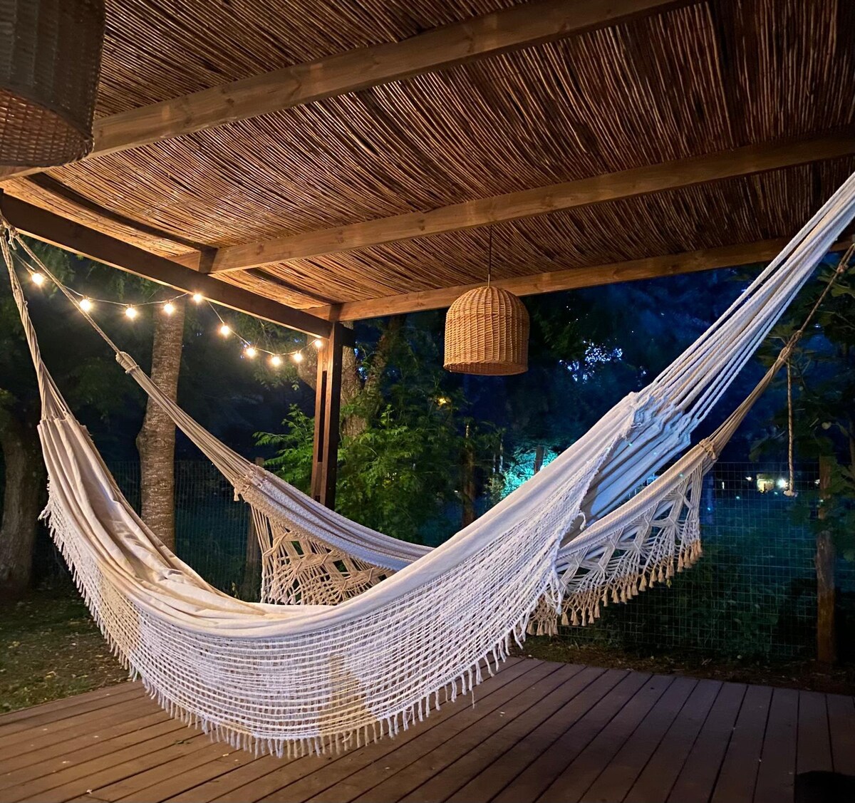 A spacious outdoor area features two white hammocks draped under a wooden pergola. Soft lighting is provided by hanging bulbs, adding a warm ambiance. Lush greenery is visible in the background, creating a serene environment for relaxation.