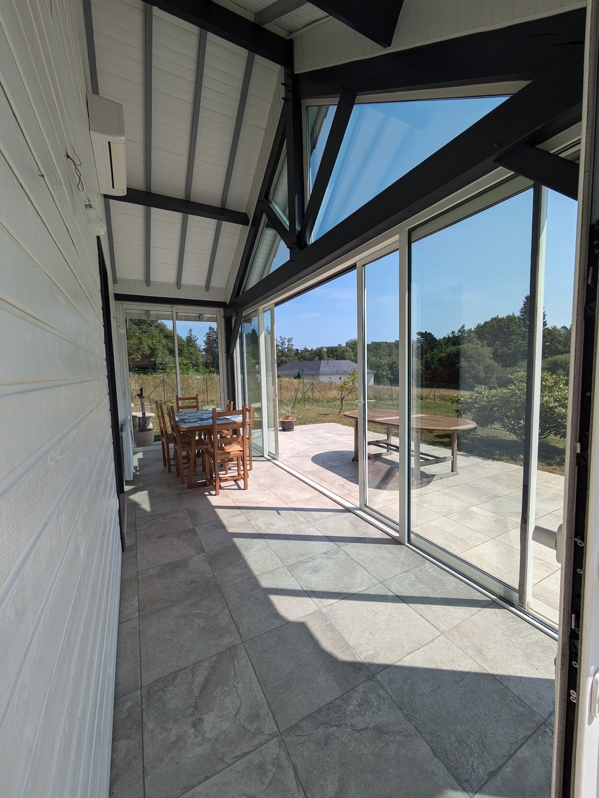A sunlit terrace is depicted, featuring a dining table surrounded by chairs. Large glass panels create an open view towards the surrounding landscape, allowing natural light to fill the space. The flooring combines tiles and wood, enhancing the blend of indoor and outdoor areas.
