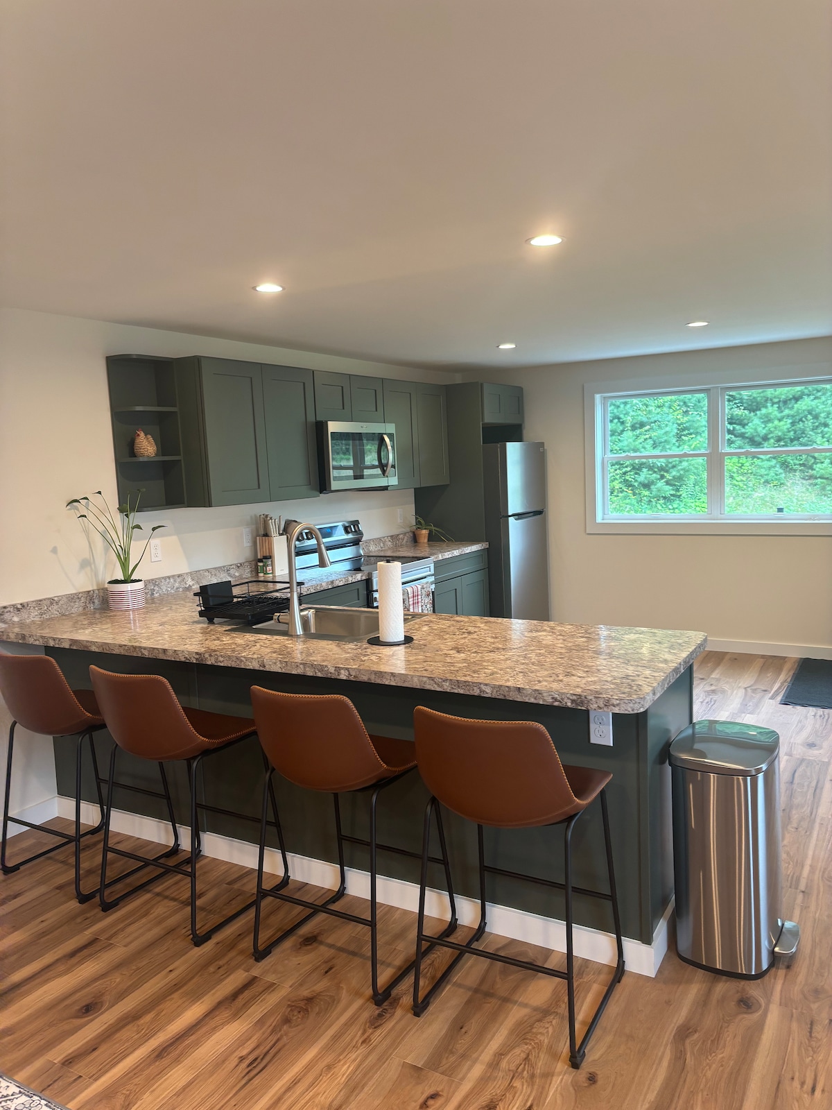 The modern kitchen features dark green cabinetry and a large granite countertop with seating for four. Stainless steel appliances are included, and a window provides natural light, illuminating the open space. A trash can is situated next to the counter for convenience.