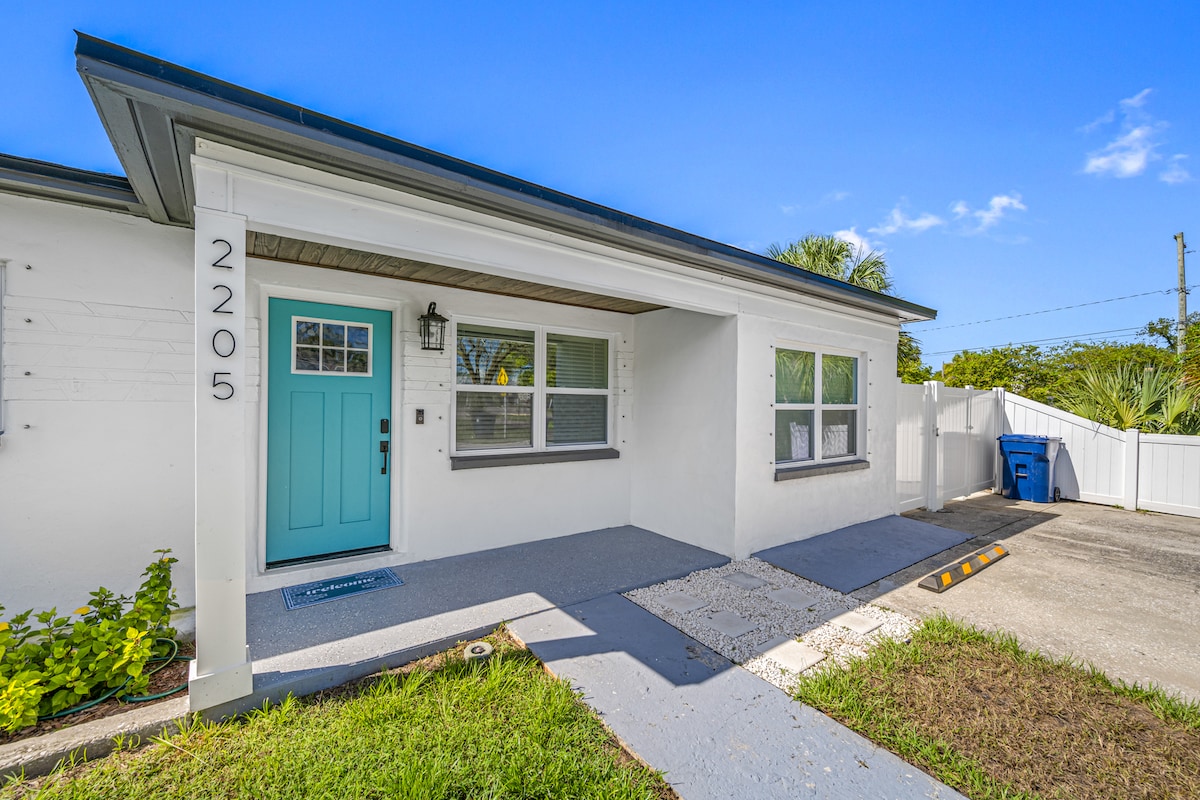 The exterior of the property showcases a welcoming entrance with a light blue door, flanked by two windows. A concrete pathway leads to the entrance, surrounded by well-maintained landscaping. A parking space is visible on the side, with the driveway extending towards a fence.