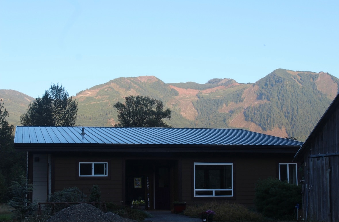 A single-story retreat is framed by majestic mountains in the background. The structure features a metal roof and several windows that allow natural light to enter. A garden area is visible in the foreground, adding to the serene environment.