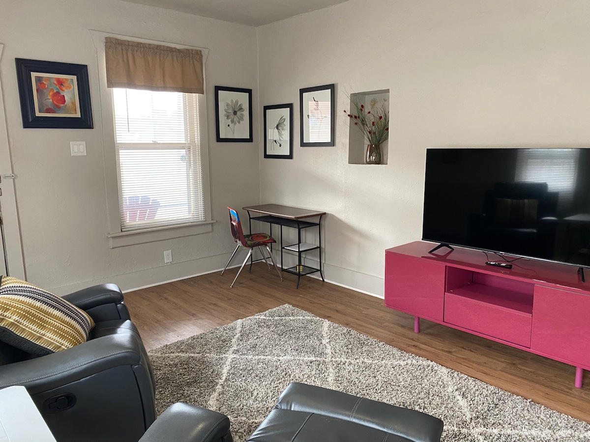 A living area features a contemporary pink media console with a flat-screen television. A small desk with a chair is positioned beside a window adorned with natural light. Framed art pieces and soft carpeting add texture to the space, complemented by seating options.