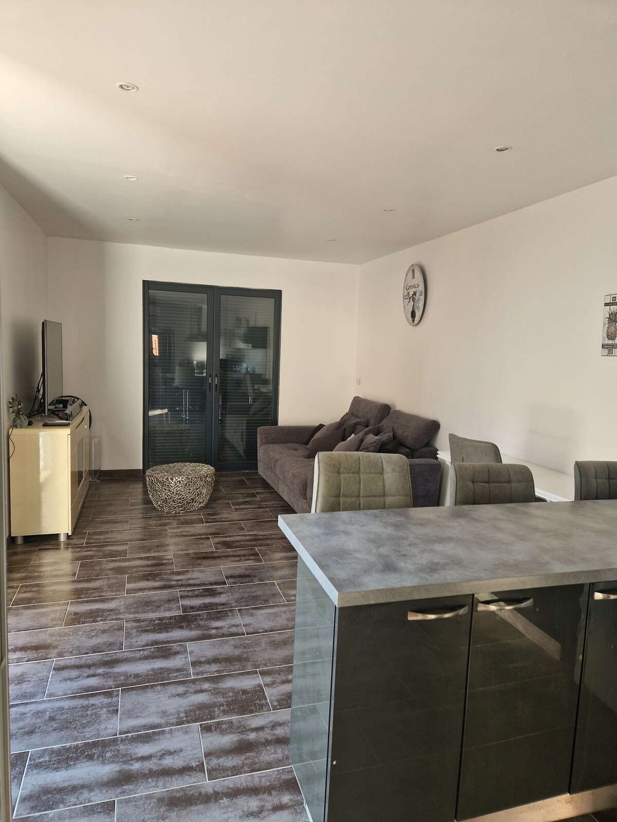 A spacious living area features a large gray sofa facing a wall-mounted television. A dining table with chairs is positioned nearby, complementing the sleek kitchen cabinets. Natural light illuminates the tiled floor, enhancing the open layout.