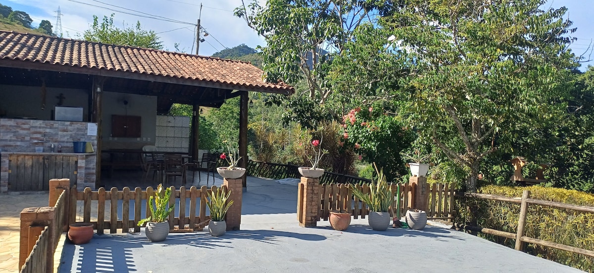 An outdoor area is featured with a spacious cement surface, surrounded by a wooden fence. Potted plants are arranged along the edge, and a covered structure with a tiled roof is visible in the background. Lush greenery and distant hills provide a natural backdrop.