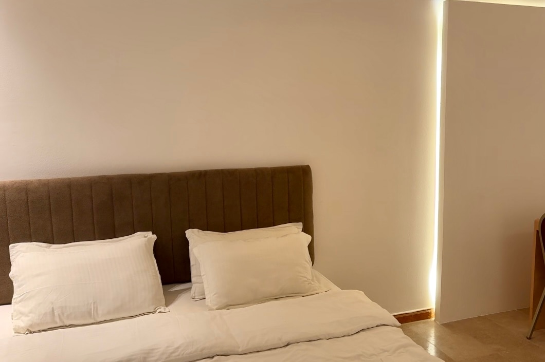 A sleeping area features a spacious bed with plump pillows and a neutral-toned headboard. Soft lighting enhances the ambiance from a nearby wall. A simple chair is positioned in the corner, providing additional seating.
