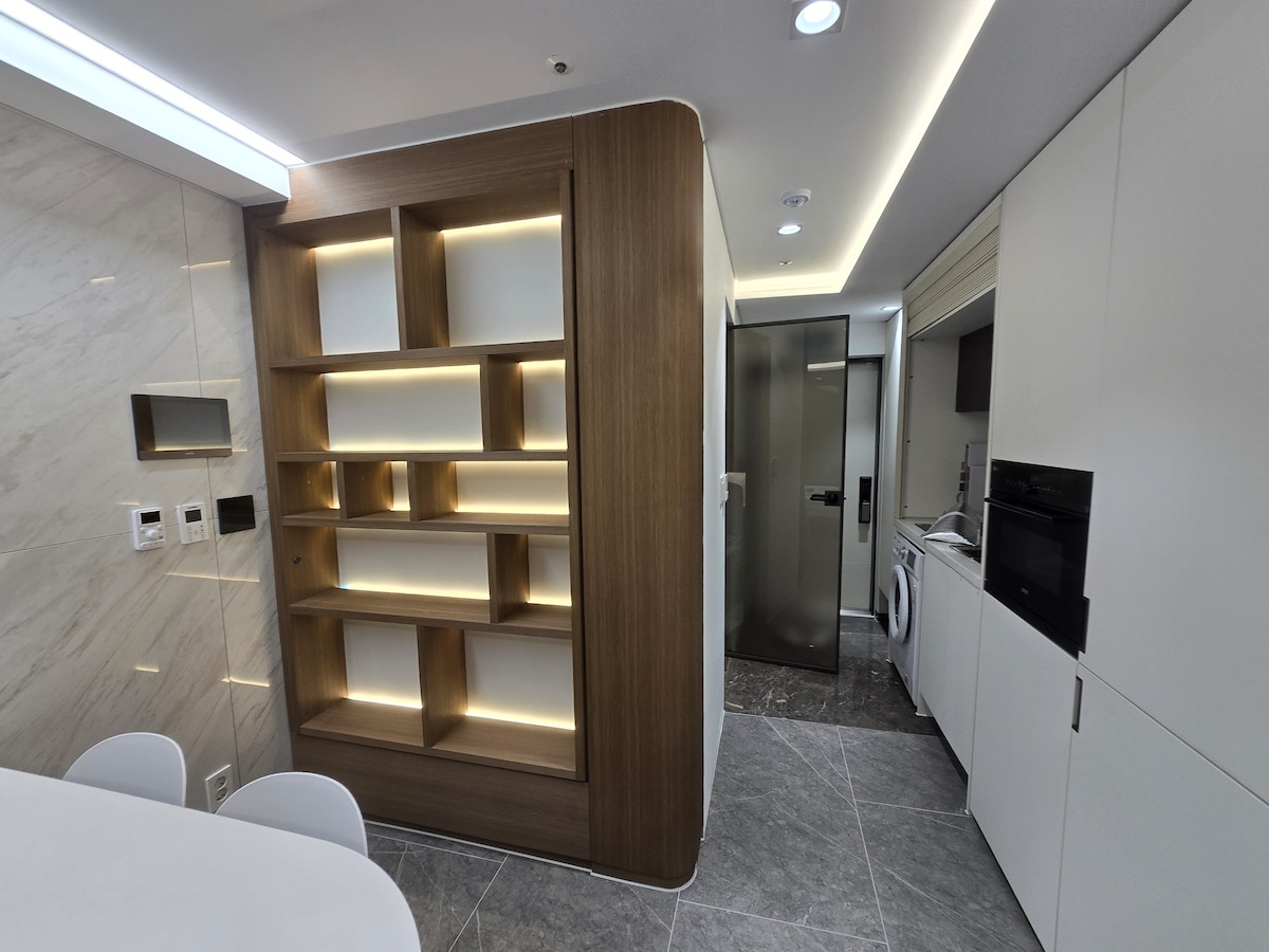 A modern kitchen area features sleek white cabinetry and a built-in microwave. A stylish wooden shelving unit is illuminated with soft lighting, providing a functional yet decorative display. Grey floor tiles and recessed ceiling lights contribute to the contemporary design.