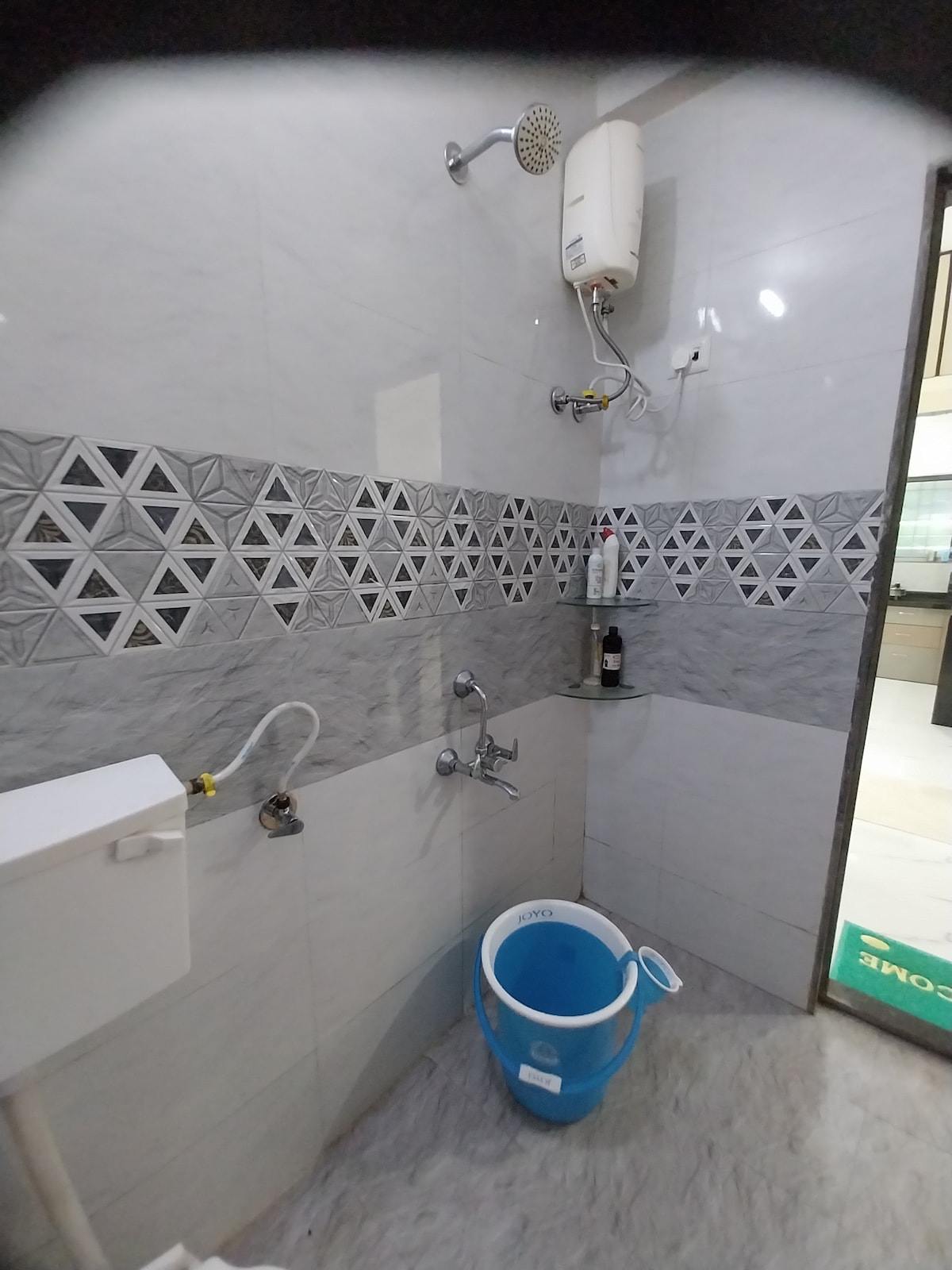 A modern bathroom is presented, featuring a geometric tile design in gray and white. A shower head is mounted on the wall above a bathing area equipped with both a handheld shower and a faucet. A clean blue bucket is placed on the floor, complemented by functional bathroom fixtures.