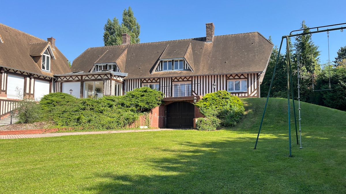 A spacious and charming building with a traditional design is set against a backdrop of lush greenery. The well-kept lawn features a children's swing set, adding a playful element to the serene environment. The structure showcases a combination of light and dark wood accents.
