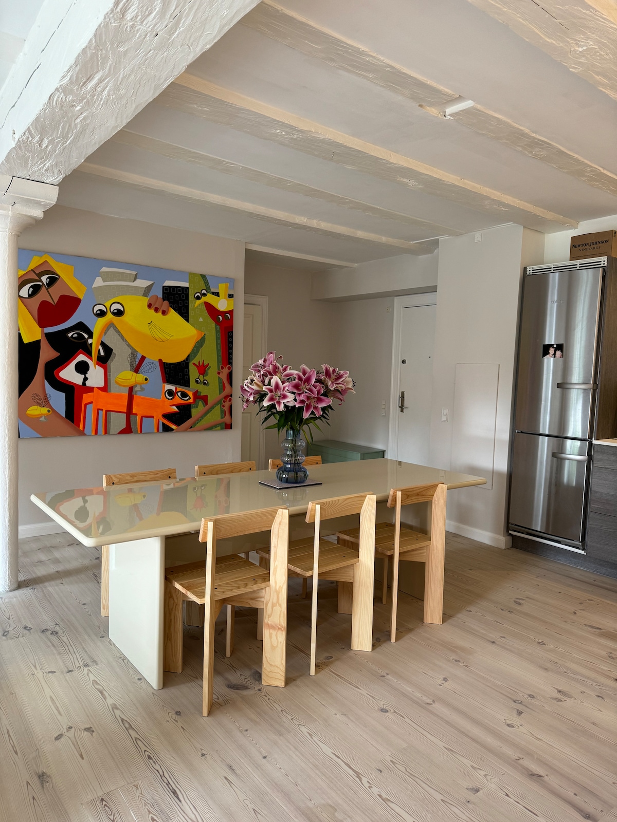 A modern dining area features a light wooden table accompanied by four simple chairs. A vibrant wall art piece adds color to the space, while a vase filled with pink lilies sits at the center of the table. Neutral tones and natural light enhance the inviting ambiance.