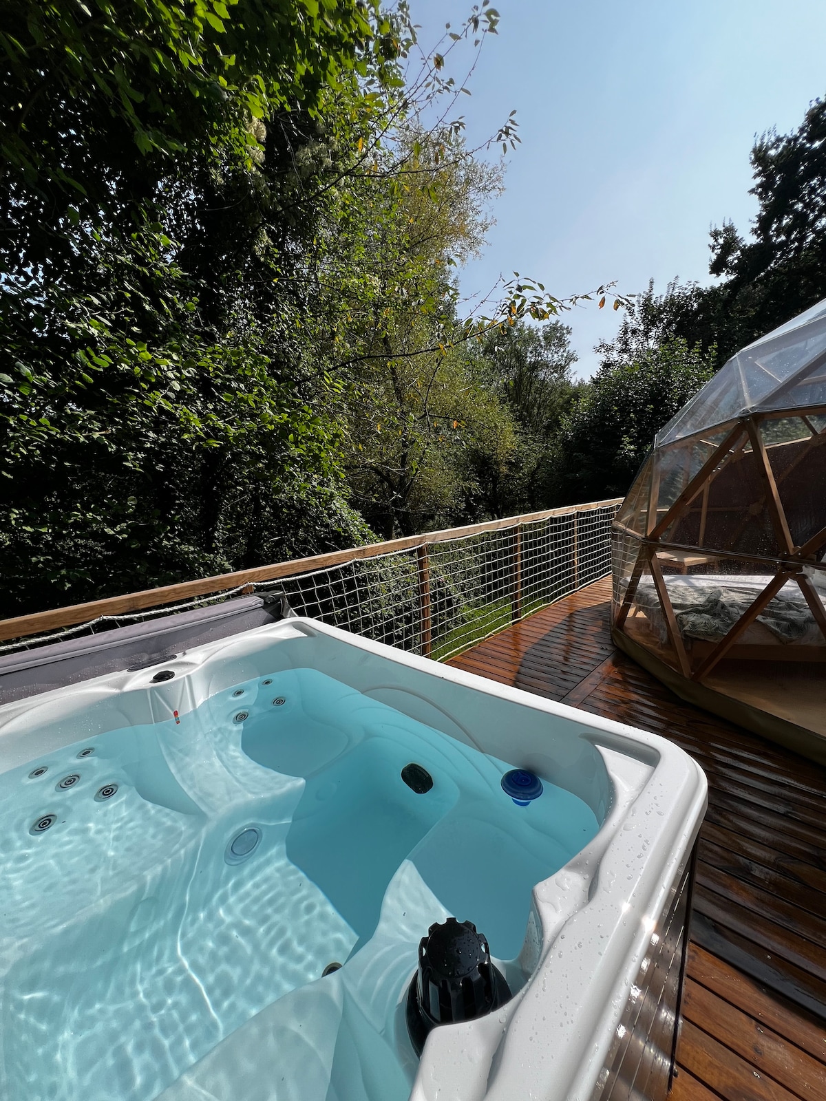 A private jacuzzi is positioned on the wooden deck, surrounded by lush greenery. Clear water reflects the sunlight, with trees providing a natural backdrop. The geodesic dome is visible in the distance, enhancing the tranquil outdoor space.