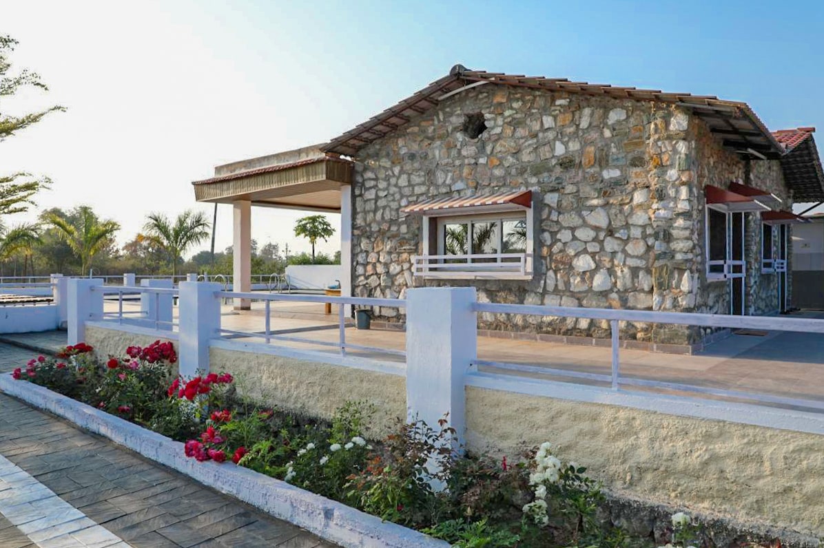 A rustic farmhouse constructed of natural stone features a sloping roof and wooden accents. Surrounding the building, lush greenery and colorful flower beds create a vibrant landscape. A spacious porch offers a welcoming entrance, while large windows allow natural light to enhance the interior.