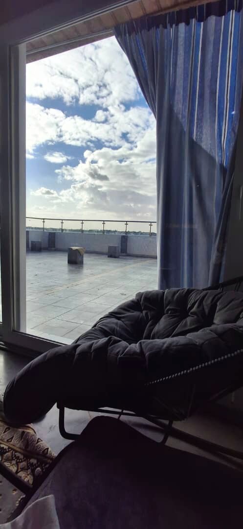 A spacious terrace is visible through large glass doors, allowing natural light to fill the room. A comfortable, dark-colored chair sits prominently, while striped curtains frame the view of the outdoor area, which features a tiled surface and a partly cloudy sky.