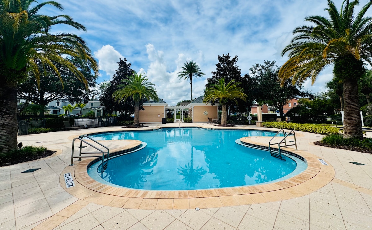 A serene pool area is surrounded by lush palm trees, providing a tropical atmosphere. The clear blue water is framed by tiled decking featuring curved edges and handrails. Comfortable loungers and landscaped greenery can be observed in the background, contributing to a relaxing outdoor environment.