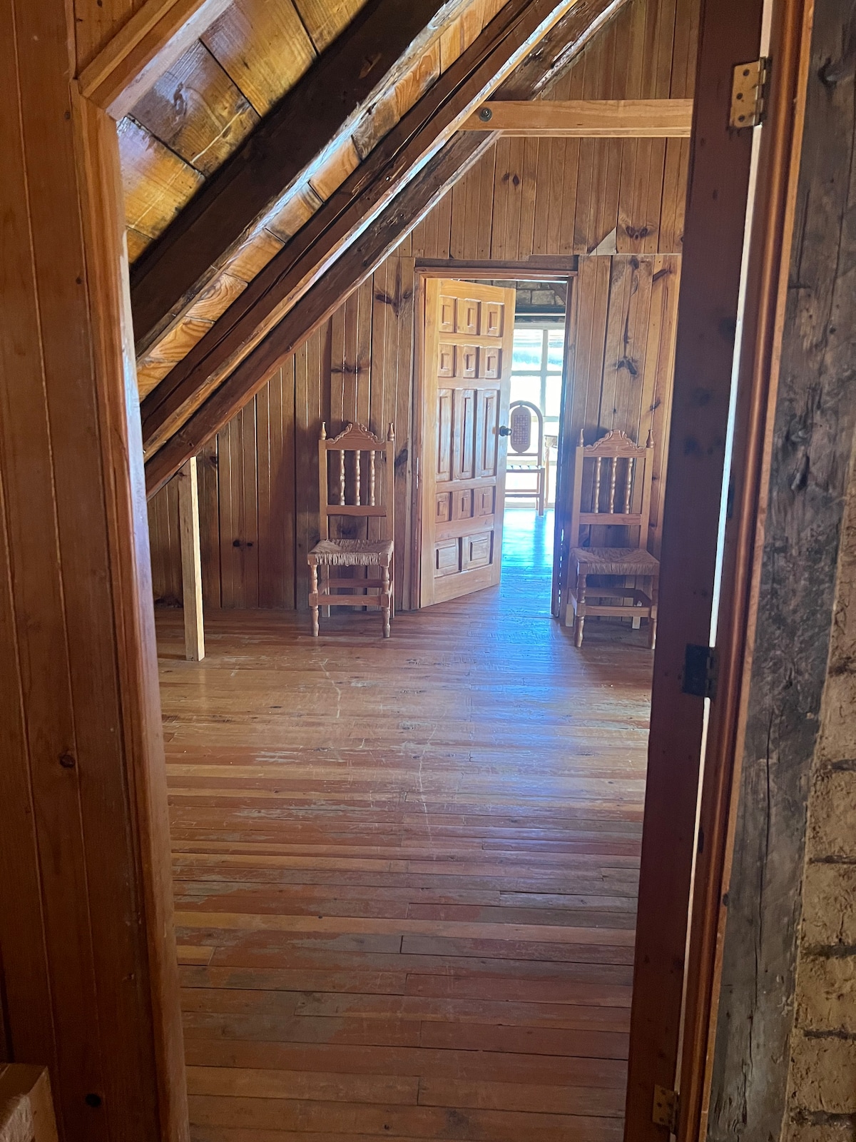 A doorway frames a view into a room featuring sloped wooden ceilings and floors. Two wooden chairs are positioned against the wall, while a second door opens to a sunlit area beyond, enhancing the sense of space.