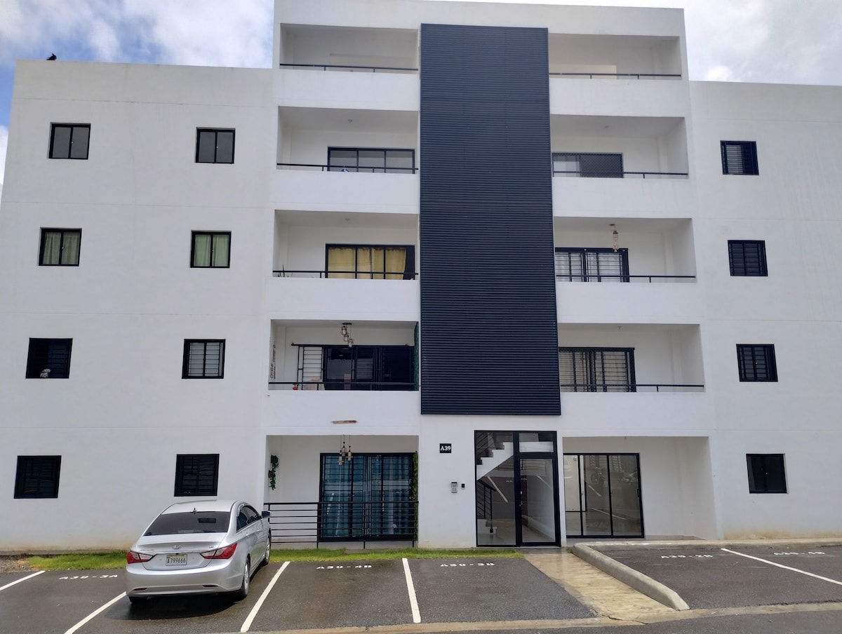 A modern three-story building features a clean white exterior with a dark accent panel. Large windows are positioned throughout, allowing natural light to enter. A sleek entryway is visible, with a parking space for a vehicle in front.