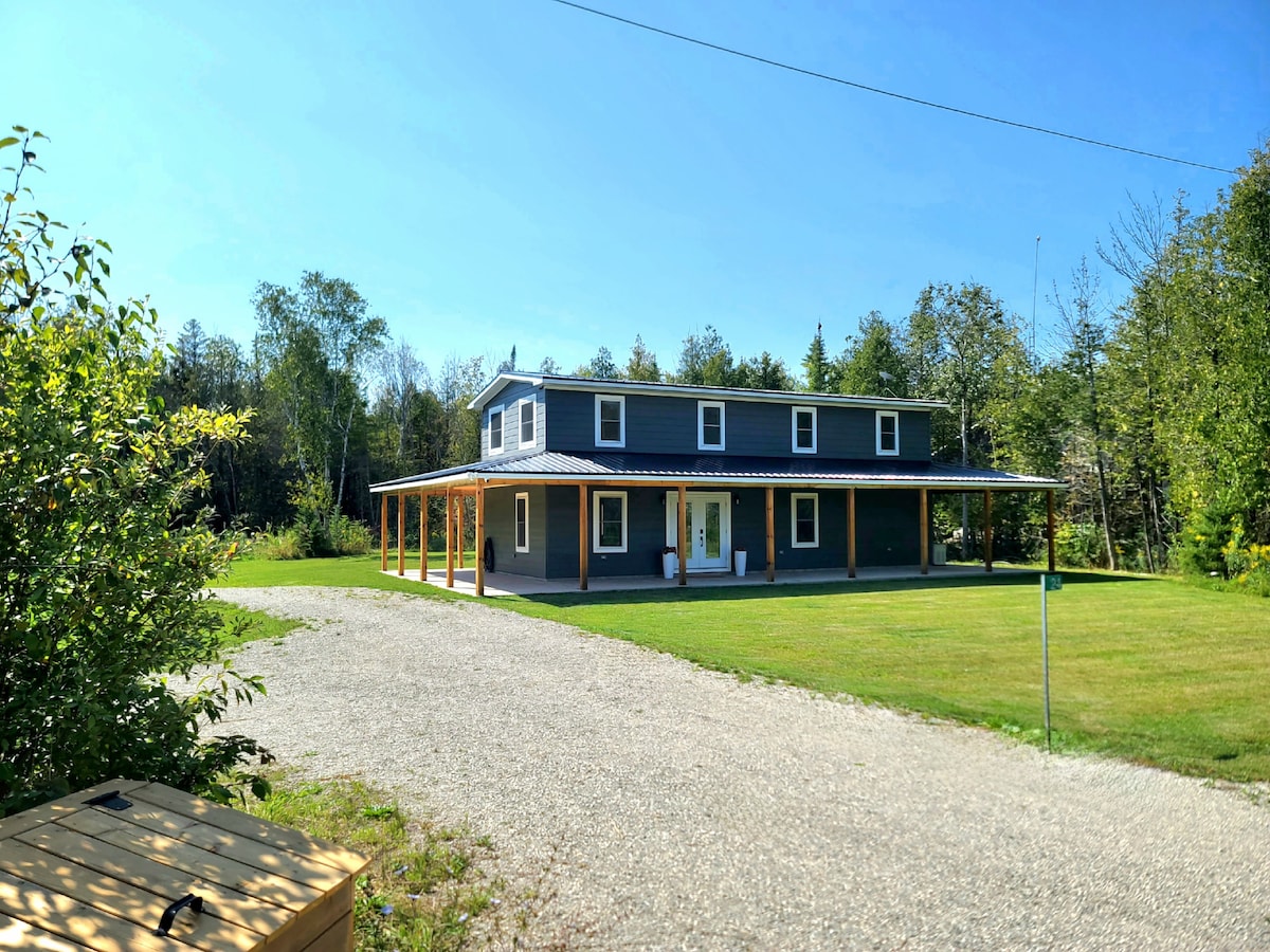 A spacious cottage is set against a clear blue sky, featuring a covered porch that wraps around the front. A neatly maintained lawn surrounds the cottage, providing ample outdoor space. The gravel driveway leads up to the entrance, bordered by patches of greenery.