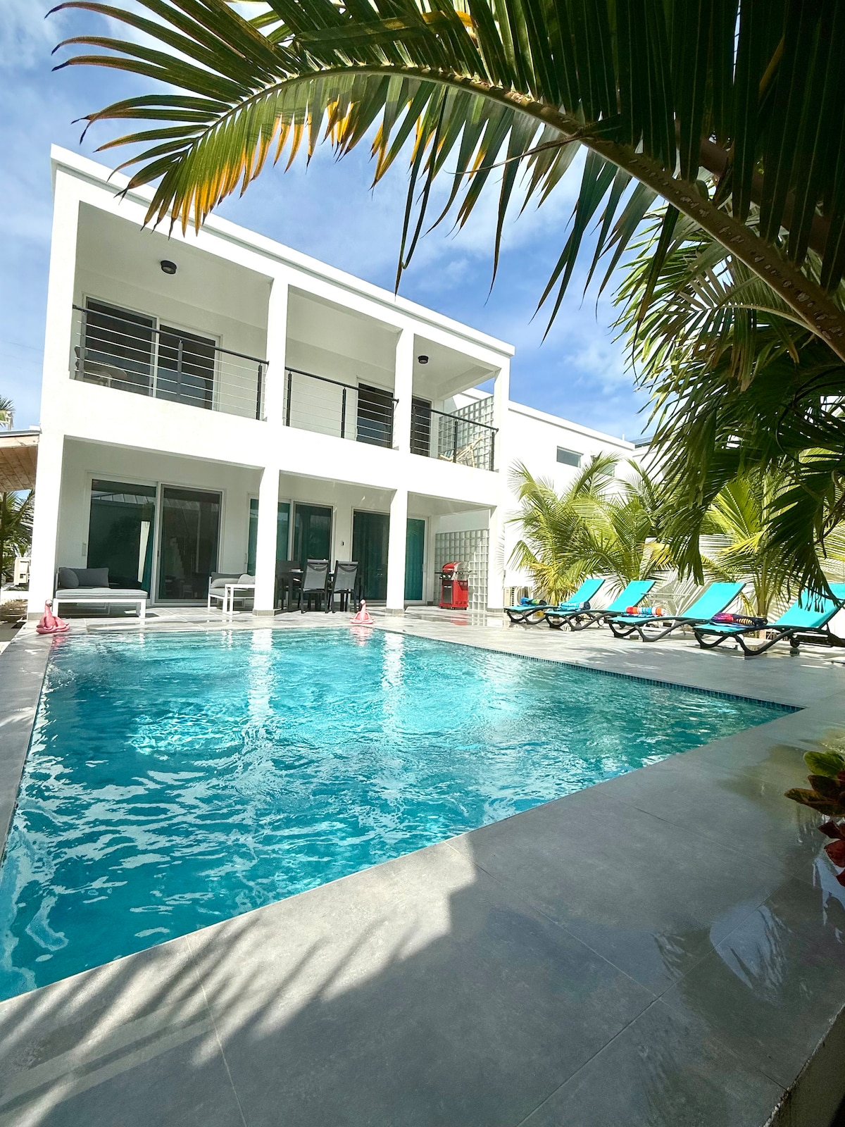 The exterior of the villa is highlighted by a private pool, surrounded by lounge chairs under palm trees. The two-story structure features large glass doors, offering views of the pool area and landscaped surroundings.