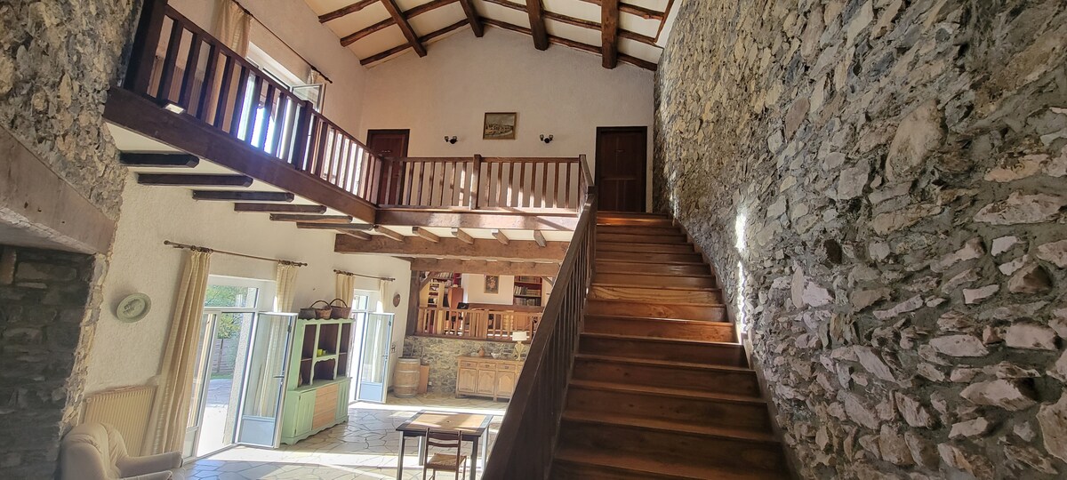 An open space is characterized by a high wooden ceiling and exposed stone walls. The wooden staircase leads to a loft area, while large windows allow natural light to fill the room. A welcoming seating area is visible, enhancing the sense of spaciousness.