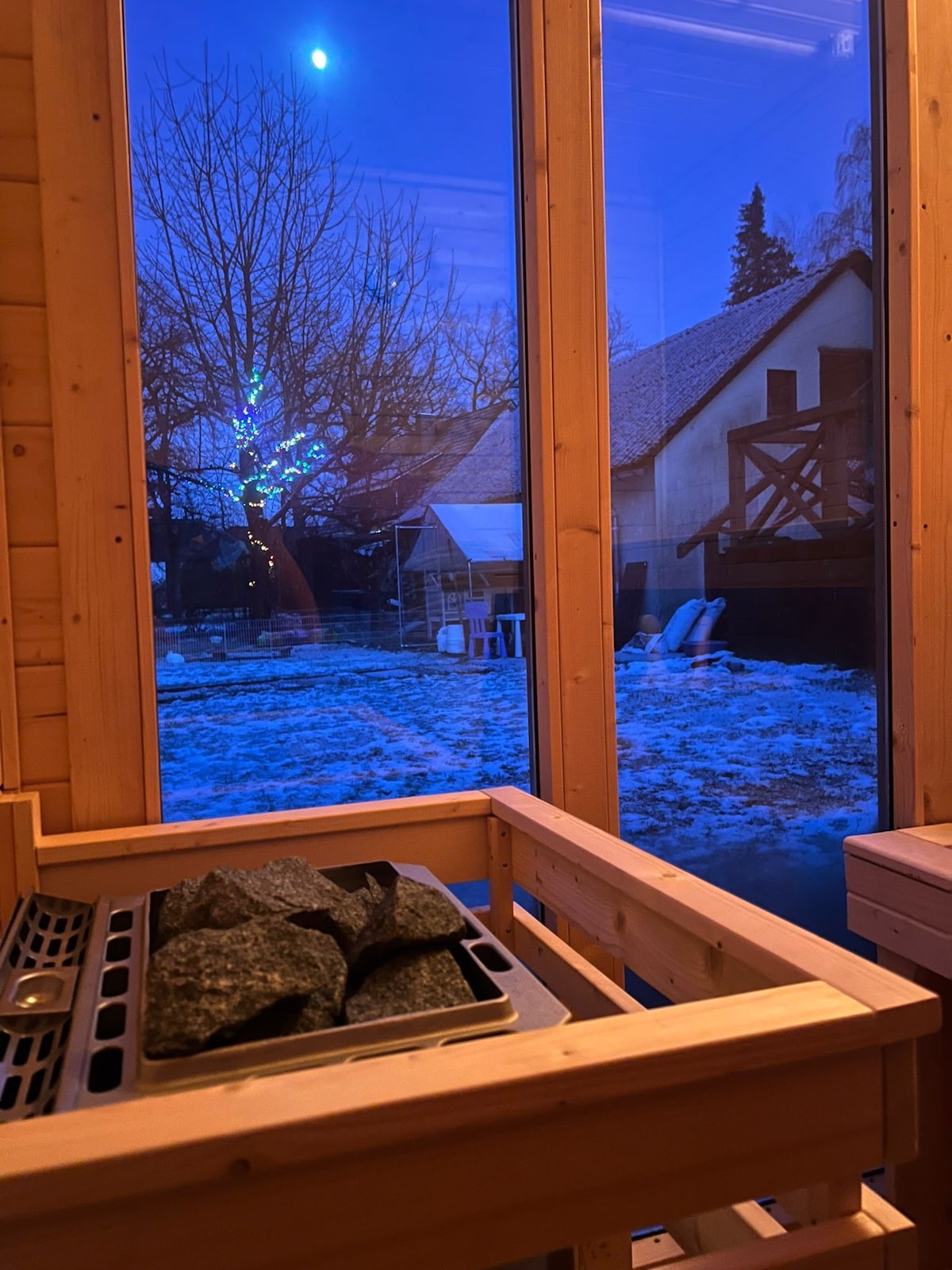 A view from inside the sauna features a wooden interior with a stone heater, overlooking a snowy garden scene. Large glass panels frame the outdoor space, where a tree is illuminated by soft blue light, highlighting a tranquil winter atmosphere.