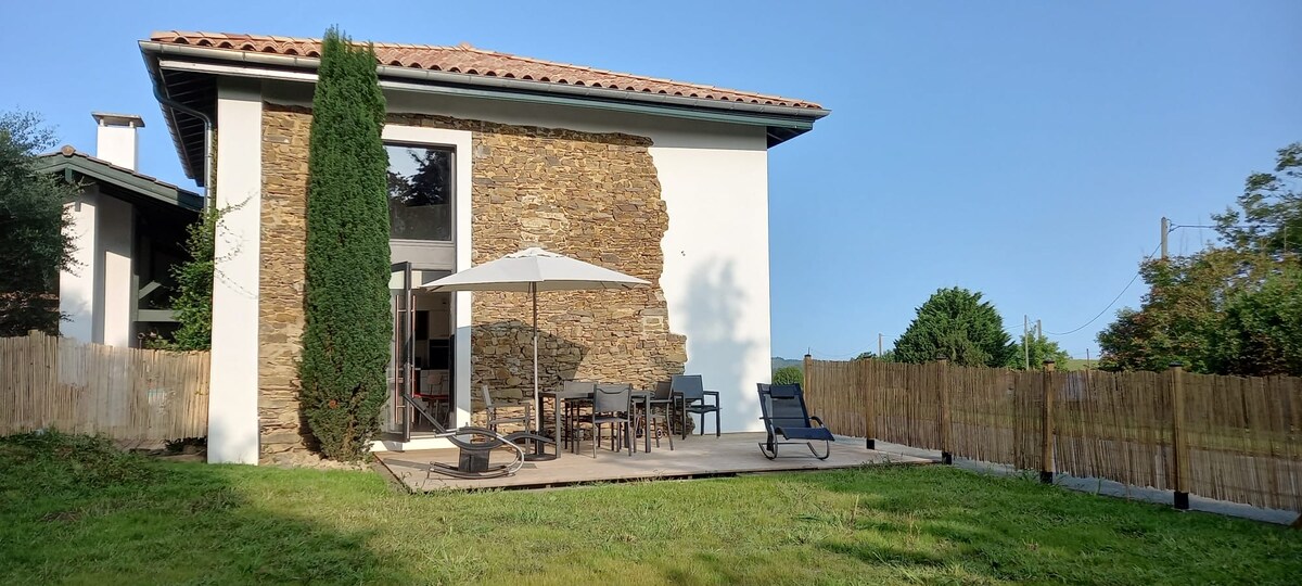 A spacious outdoor area features a wooden deck with a large umbrella providing shade for the dining table and chairs. Lounge chairs are positioned nearby on a well-kept lawn, framed by natural stone walls and a privacy fence. Clear skies enhance the tranquil setting.