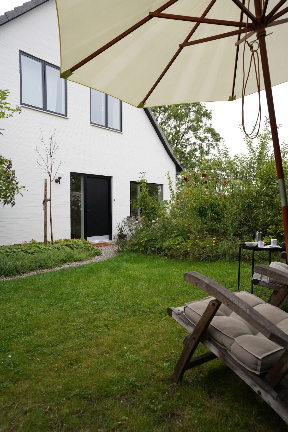 A serene garden area is presented, featuring a wooden lounge chair with cushions under a large, light-colored umbrella. The background reveals a two-story house with black windows and a black door, surrounded by lush greenery and flowering plants.